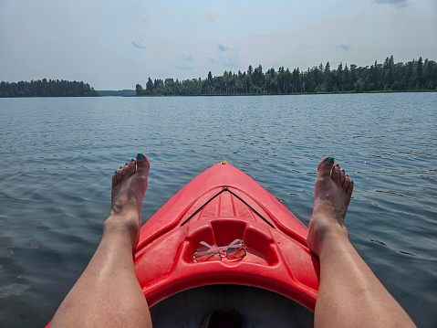 Outdoor recreational pursuit image of the bow of a red kayak floating on the water surface of a freshwater lake while the person taking the photo rests their legs on top of the kayak.