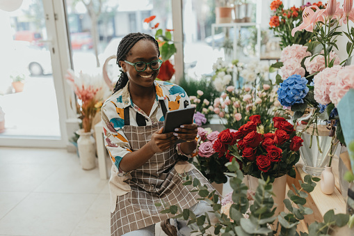 A compelling portrait featuring a talented 25-29-year-old African-American florist, proudly owning her flower shop. With a warm smile, she sits amidst a colorful array of flowers, utilizing a digital tablet for quality control and inventory management. The image beautifully blends entrepreneurship, technology, and the artistry of floral arrangements in the realm of small business.
