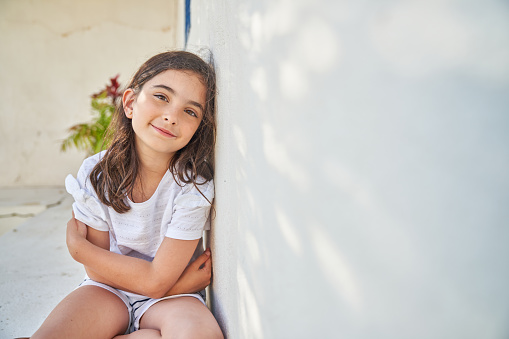 Cute smiling preteen girl with long brown hair in casual clothes sitting on sidewalk with crossed arms and leaning on concrete wall while looking at camera