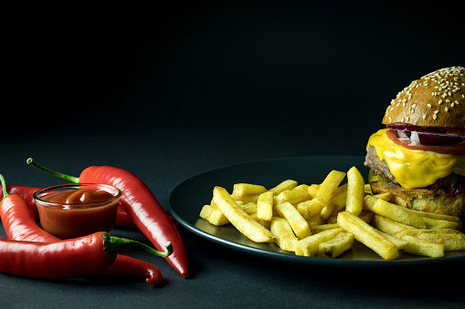 Chili peppers, spicy ketchup and burger with french fries on black background