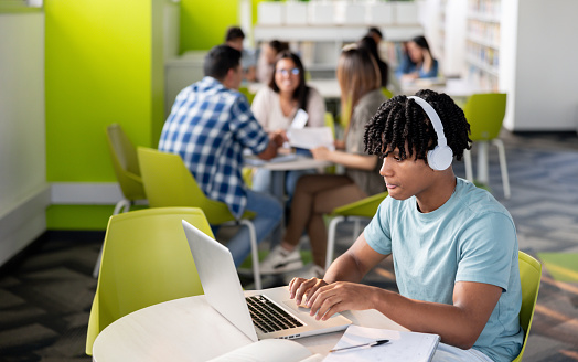 African American student studying at a public library using a laptop and headphones - education concepts