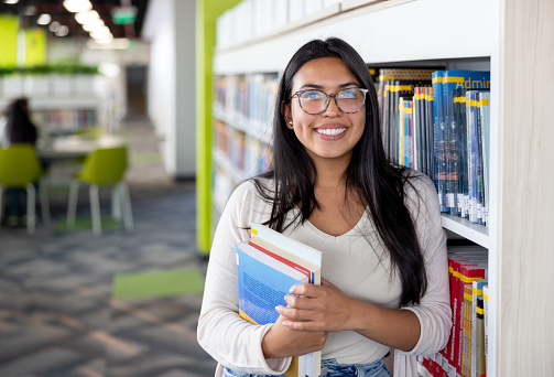 Portrait of a happy student holding books at the library and looking at the camera smiling - education concepts