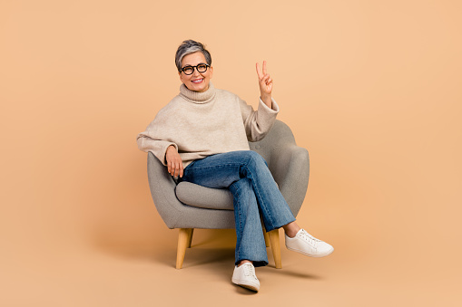 Full length photo of friendly mature woman in comfortable place armchair make peace v sign at therapy isolated on beige color background.