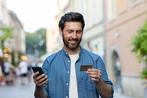 A smiling mature man with a beard uses a smartphone and credit card on a bustling city street as evening sets in.