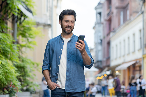 Joyful mature bearded man strolling through an urban setting at dusk, engaged with his smartphone, embodying leisure and connectivity.