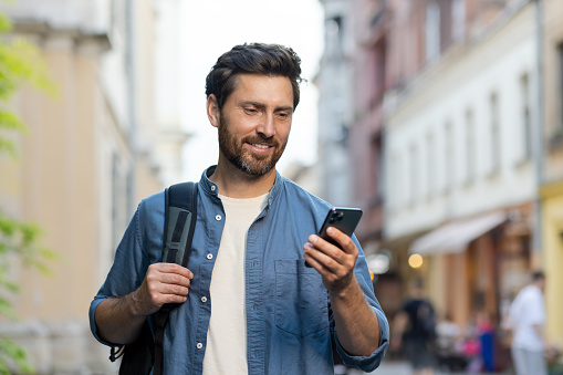 A handsome smiling young man wearing a denim shirt and carrying a backpack is standing on a city street and using a mobile phone.