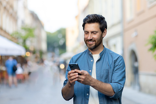 A smiling young man is standing on a city street in a denim shirt and using a mobile phone. Close-up photo.