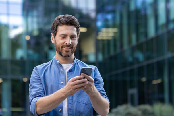 istock Smiling businessman with beard using smartphone outside modern office 2030401595