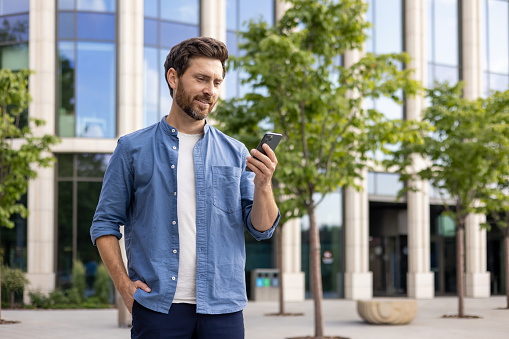 A smiling young man in casual clothes is standing on a city street near a skyscraper, keeping his hand in his pocket and looking at the phone screen.