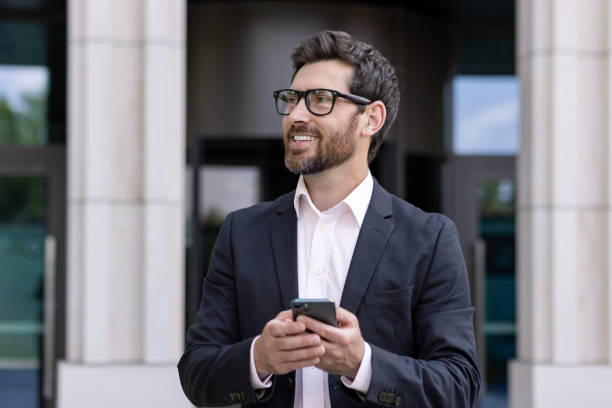 istock Confident businessman with beard using smartphone outside office building in city 2030421174