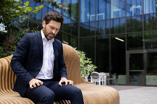 A young male businessman is sitting in a suit on the street outside an office center on a bench and is suffering from itchy body, suffering from severe stress and panic attack.