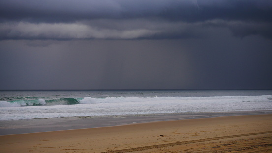 View of a storm on the blue emerald sea, horizon on water and shore with white surf on the waves, cloudy dark sky with rain in the background