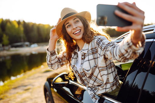 Happy woman on vacation taking selfie photo with smartphone near car. Stylish model on a car trip. Travel, blogging concept.