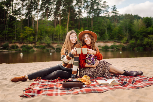 Two young women making picnic at the beach. Positive models sitting on a red mat with a thermos and coffee. Females enjoys nature at mini picnic. Concept of relaxation, nature.