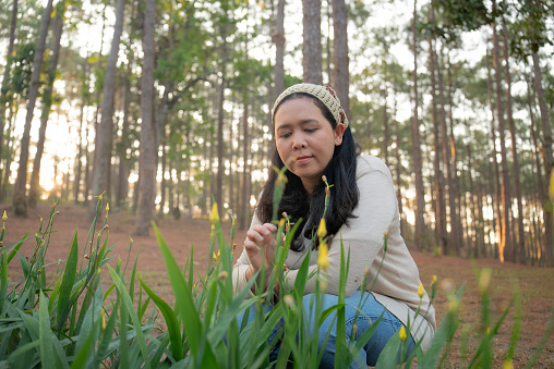 Happy Asian mid-adult woman is looking the flowers on the ground in the forest, feeling nature with a positive and refreshing mood