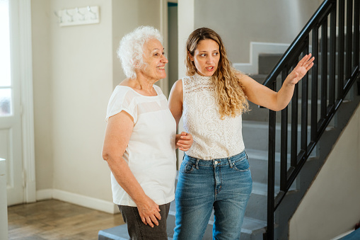 Young adult woman guiding a happy senior woman through a home interior, expressing care and assistance.