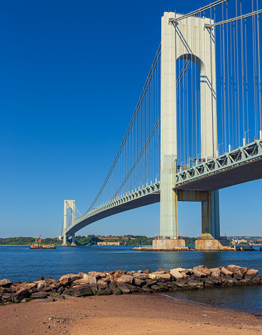 Verrazano-Narrows Bridge on a Summer Morning with Water of New York Harbor. The bridge connects boroughs of Brooklyn and Staten Island in New York City. It was built in 1964 and is the largest suspension bridge in the USA. Historic Fort Wadsworth is next to the foot of the bridge. Industrial Boat passing under the Bridge. Canon EOS 6D full frame censor camera. Canon EF 24-105mm f/4L lens.