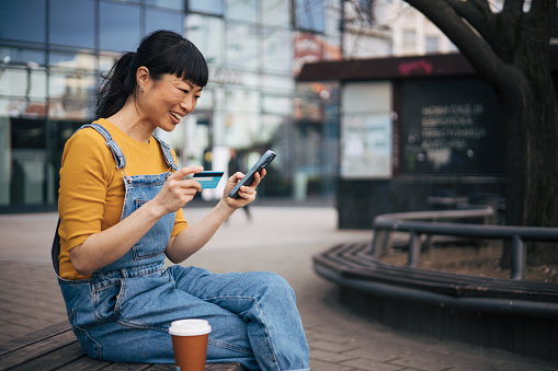 A Yapanese woman sits on a bench and using credit card for online shopping.She drinks takeaway coffee and laugh