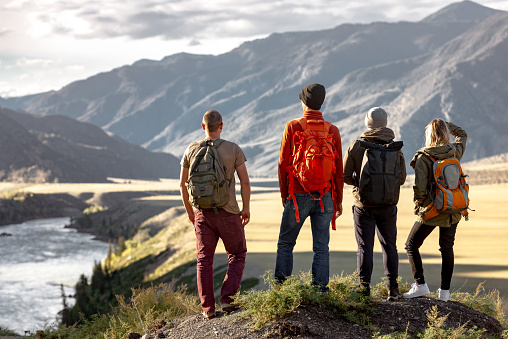 Group of young hikers with backpacks are standing at observation point in mountains and enjoying nice view at sunset and big river