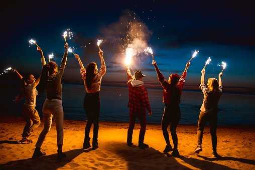 Big group of happy young friends are celebrating something and having fun with signal fire at sunset lake beach