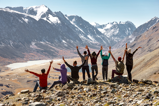 Big group of happy young friends or tourists are standing and sitting with open arms high in mountains