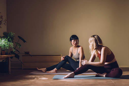 A peaceful scene of two women in yoga attire engaging in a friendly chat while sitting on mats in a well-lit room.