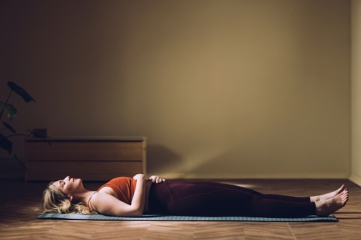 Relaxed female in sportswear lying in Corpse Pose, mindfulness meditation, calm indoor setting, well-being concept.