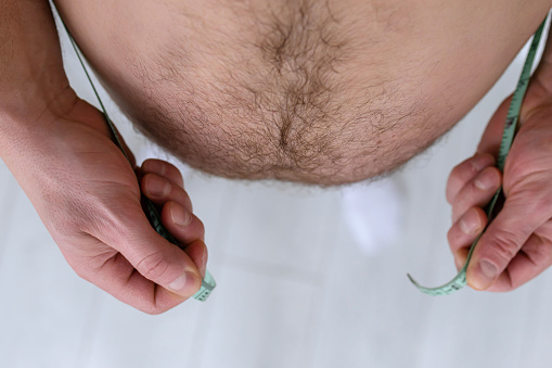 Male measuring his belly with a centimeter. Man with bare fat belly shakes fat folds on his stomach, obesity, health, beer belly. Problem area of body. Hormones. Overweight. Copy space