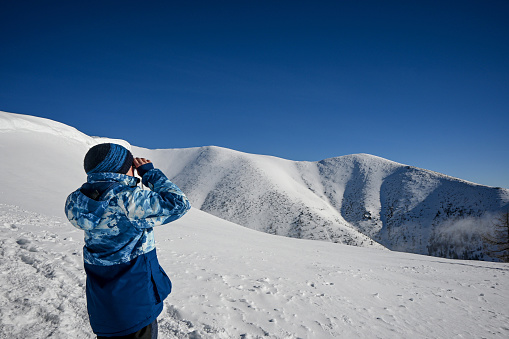 A 10-year-old boy is looking through binoculars at a beautiful snow-covered mountain in the western Tatras of Baranec, the weather is winter and beautiful with a blue sky.