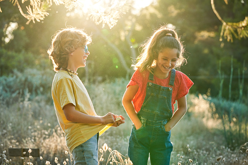 Happy boy and girl with hands in pockets telling jokes while standing in field with dry grass together on sunny summer day in countryside