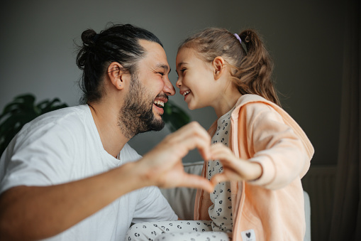 Head shot portrait of smiling father with cute daughter showing heart, looking at camera, happy dad and little child posing for funny family photo in living room, sitting on couch together. Dad and girl kid make hand heart gesture. Happy Father's day.