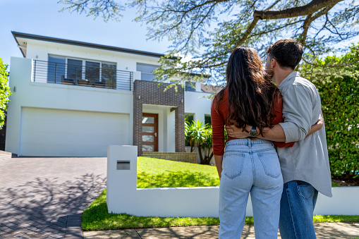 Happy Couple standing in front of their new home. They are both wearing casual clothes and embracing. Rear view. The house is contemporary with a brick facade, driveway, balcony and a green lawn. The front door is also visible.