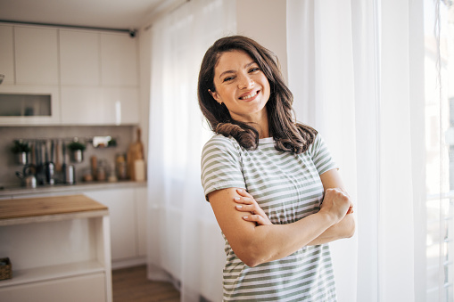 Portrait of smiling young woman with arms crossed standing in the kitchen  behind the window. She is looking at camera.