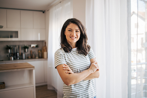 Portrait of smiling young woman with arms crossed standing in the kitchen  behind the window. She is looking at camera.