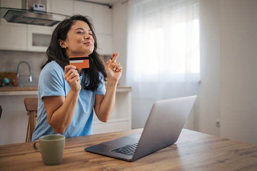 Woman with credit card shopping online through laptop at home. She is sitting at the kitchen desk. Hoping for her item she wants is available.
