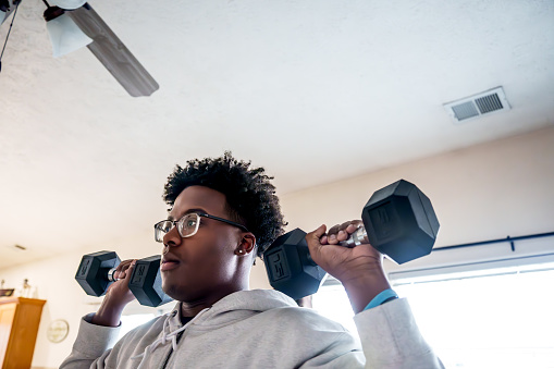 Young Determined African American Man in His Twenties Standing and Doing Curls and Military Press in a Middle Class Living Room, Home Fitness