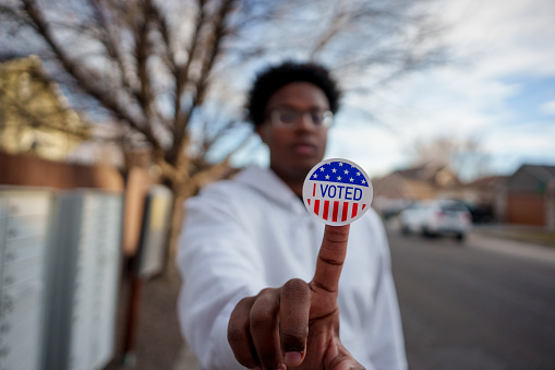 Young African American Man in His Twenties Holding an I Voted Sticker Up to the Camera and Smiling