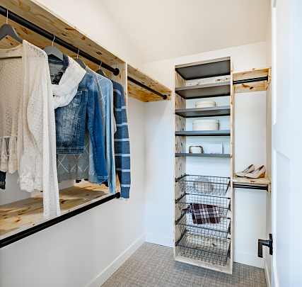 Interior view of a walk in closet with wire baskets and pine shelving. There is a denim jacket on a hanger and a white scarf. Shoes and blouse along with some decor items on the shelf.