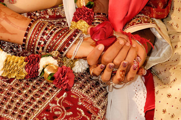 Close-up of a woman s hand, adorned with intricate henna designs and wearing gold bangles, placing a ring on a man s finger. She s dressed in a vibrant pink outfit embellished with sequins. The man s hand is brown, wearing a black suit and a simple gold bracelet. The scene captures a ceremonial moment, likely part of a cultural or matrimonial tradition.