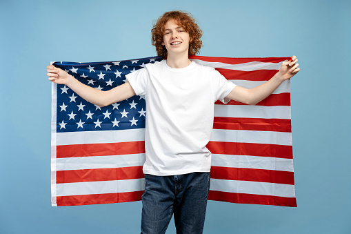 Picture of a happy, curly haired redhead teenage boy wearing braces, in casual clothing, holding the USA flag behind his back, isolated on a blue background. Emphasizes of a student visa concept
