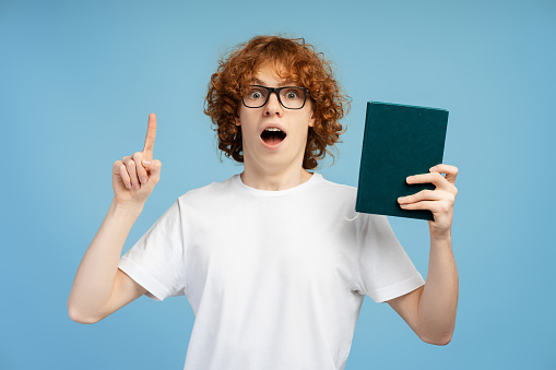 Portrait of excited curly haired redhead teen with braces and glasses, demonstrating a book and pointing upwards, making index finder gesture, isolated on blue background. Embodying study nerd concept