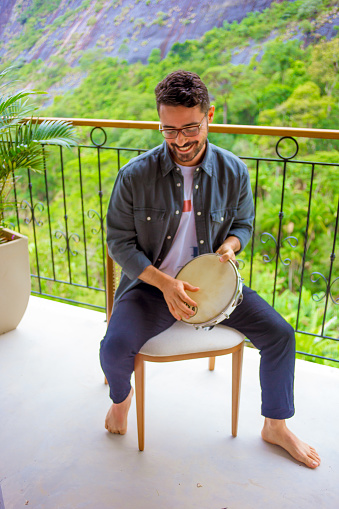 man smiles and plays the tambourine on the hotel balcony, wearing a shirt and pants and is barefoot