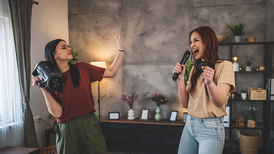 Two women young caucasian friends or sisters have fun at home females dance and sing karaoke hold microphone listen to the music happy smile joyful rhythm real people copy space