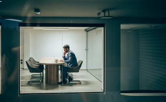 Shot of businessman sitting on conference table in the board room during night. Overworked businessman staying late hours in the office. The view is through glass.