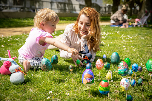 Mother and little daughter celebrate Easter in the park on a sunny spring day with colored eggs and bunnies. A young beautiful red-haired mother plays with her blonde baby. Around them are hand-made bunnies and colorful eggs painted by a little girl.