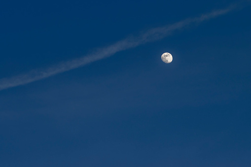 Waxing gibbous moon at twilight in a sky with faint clouds and airplane contrail (vapor trail) above the moon.