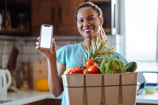 African American woman holding a cardboard box full of organic food and vegetables and showing blank smart phones screen