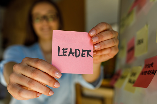 Close-up shot of a woman holding a sticky note with the word 'leader' written on it and pointing it towards the camera.