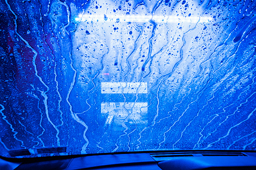 Automated car wash driver's point of view as high pressure sprayed rinsing water rapidly dilutes the car wax solution previously applied and drains in slippery twisting rivulets down the windshield glass.