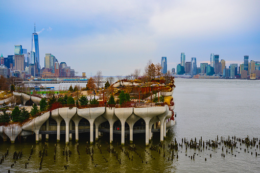 New York City and Jersey City Skyline over the Hudson River and Little Island Park Waterside Amphitheater with trees and curving foot trails on a dramatic cloudy winter evening in Manhattan, USA